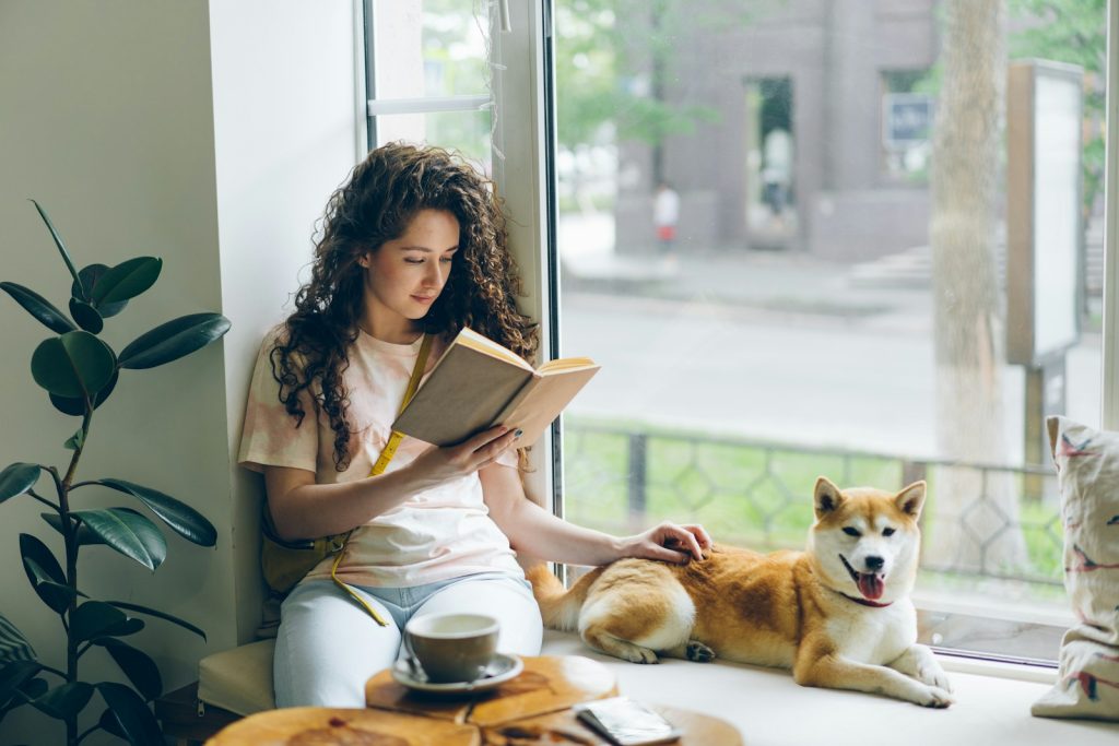 A woman sitting on a windowsill relaxing and reading a book next to two dogs